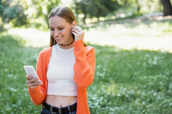 ワイヤレスイヤフォンを装着し公園内でスマートフォンを使う陽気な女性 — ストック写真