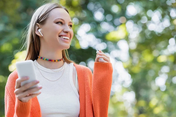ワイヤレスイヤフォンを装着し公園内でスマートフォンを使う幸せな女性 — ストック写真