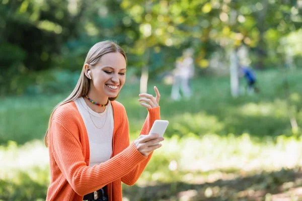 緑の公園でスマホを使いながらジェスチャーをするワイヤレスイヤフォンの陽気な女性 — ストック写真