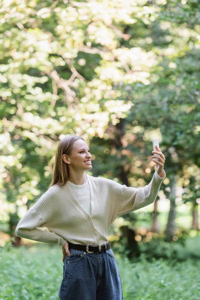 Счастливая Молодая Женщина Смартфоном Стоит Рукой Бедре Парке — стоковое фото