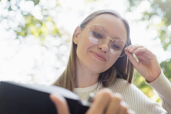 low angle view of smiling woman in glasses holding blurred notebook