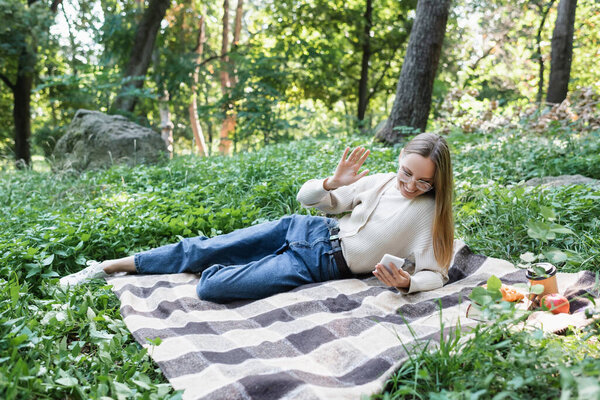 woman in glasses having video call on smartphone while lying on plaid blanket in green park