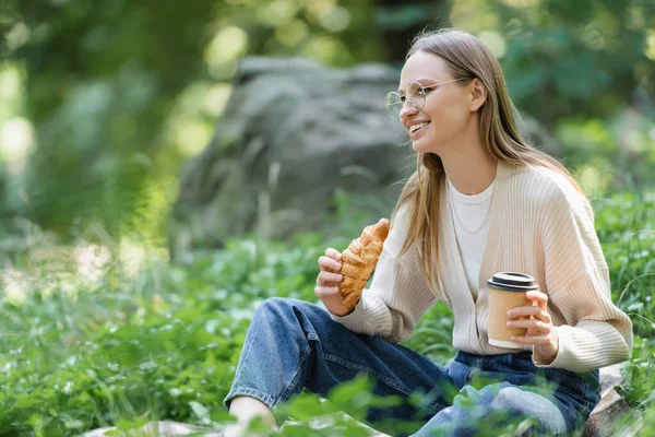 野餐时坐在草地上 戴着眼镜 手里拿着纸杯和羊角面包的快乐女人 — 图库照片