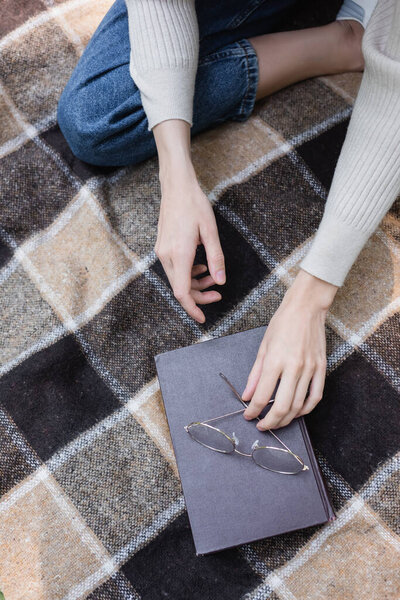 top view of woman reaching glasses on book while sitting on checkered blanket 