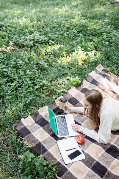 high angle view of woman in glasses lying on blanket and using laptop in park 