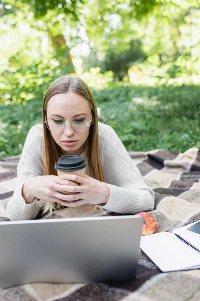 woman in glasses lying on blanket, holding paper cup and using laptop in park 