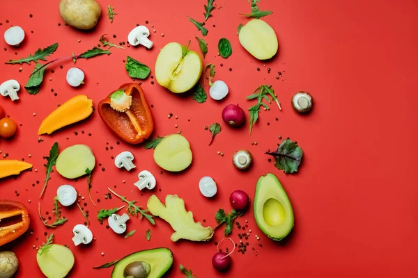 Top view of cut fruits and vegetables near peppercorns and greens on red background