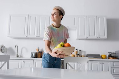 Gülümseyen kadın mutfak masasının yanında taze meyvelerle kaseyi tutarken yüzünü çeviriyor.