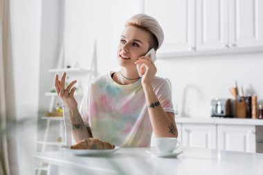tattooed woman gesturing and talking on smartphone near croissant and coffee cup
