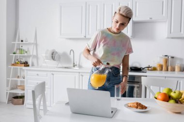 stylish woman pouring orange juice near laptop, croissant and fruits in kitchen