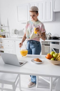 young woman with orange juice near laptop, croissant and fresh fruits on kitchen table