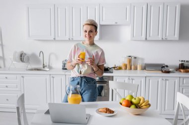 trendy woman with orange juice smiling at camera near kitchen table with laptop and breakfast
