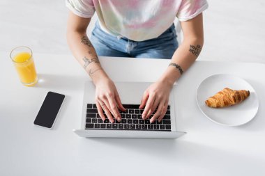 top view of cropped woman typing on laptop near orange juice, croissant and mobile phone