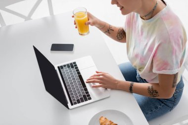 partial view of smiling tattooed woman with orange juice near laptop, smartphone and croissant