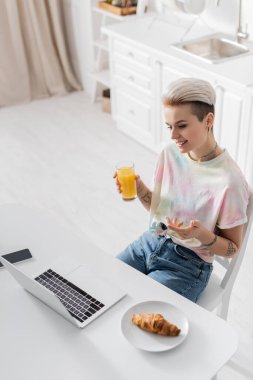 Portakal suyu ile gülümseyen kadın kruvasan ve akıllı telefonun yanındaki boş ekran bilgisayarı işaret ediyor.