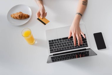 Kredi kartıyla portakal suyu, kruvasan ve cep telefonunun yanında dizüstü bilgisayarda yazan kesilmiş kadının üst görüntüsü.