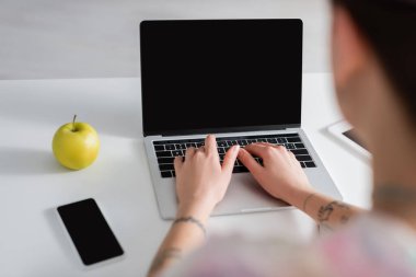 Akıllı telefonun yanında, boş ekran ve olgun elmayla bilgisayar başında yazan bulanık kadın görüntüsü.