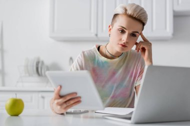 Mutfakta dizüstü bilgisayarının yanında dijital tabletli düşünceli kadın.