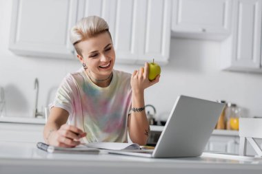 Mutfakta laptopun yanında gülümseyen olgun elmalı trend bir kadın.