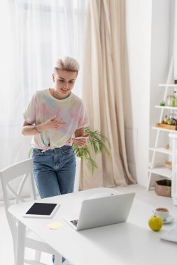 cheerful woman with smartphone pointing with hand during video call on laptop in kitchen