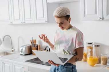 trendy tattooed woman looking at laptop and gesturing in kitchen
