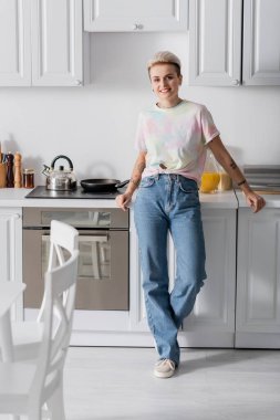 full length view of smiling woman in jeans looking at camera in kitchen