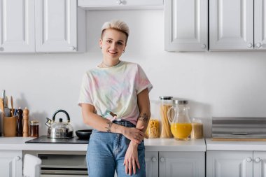 tattooed woman with trendy hairstyle smiling at camera in kitchen with white furniture