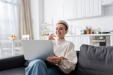 excited woman with trendy hairstyle laughing near laptop on sofa at home clipart