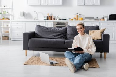 cheerful woman sitting on floor with notebook and laptop near couch in open plan kitchen clipart