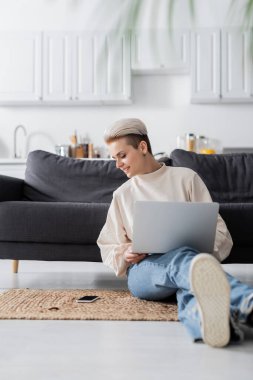 smiling woman sitting on floor with laptop and looking at smartphone with blank screen clipart