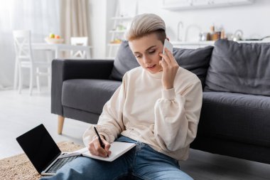 young woman talking on smartphone and writing in notebook near laptop with blank screen clipart