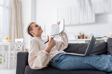 excited woman with blank notebook laughing near laptop on couch clipart