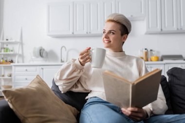joyful woman with book and cup of tea sitting on sofa at home and looking away clipart