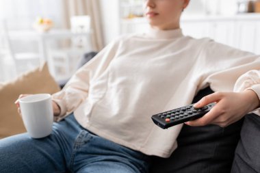 Çay fincanı ve uzaktan kumandayla evde televizyon izleyen bulanık kadının kısmi görüntüsü
