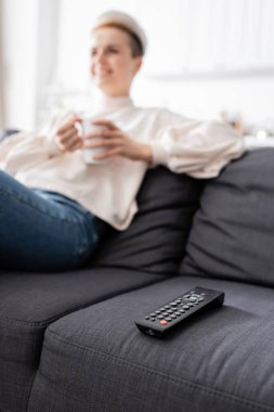 partial view of blurred woman with cup of tea sitting on couch near tv remote controller