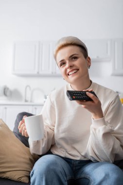 smiling woman holding cup of tea while clicking tv channels at home