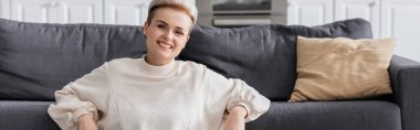 happy woman in white pullover smiling at camera near couch and pillow, banner