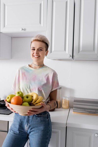 cheerful and stylish woman with bowl of ripe fruits smiling at camera in kitchen