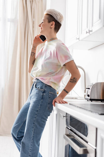 side view of happy woman in jeans standing in kitchen and talking on cellphone