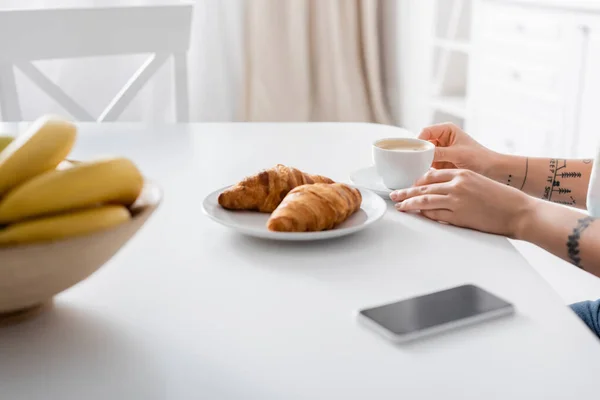 Cropped View Tattooed Woman Coffee Cup Croissants Blurred Bananas Smartphone — Stok Foto