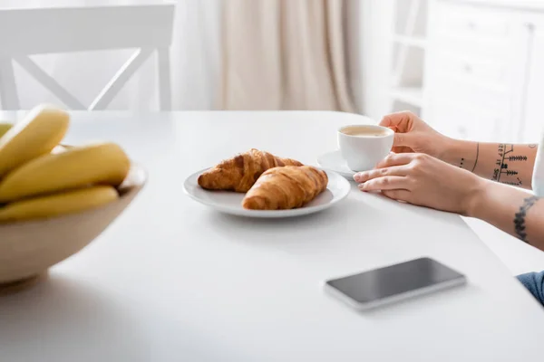 Ostříhaný Pohled Tetovanou Ženu Šálku Kávy Croissanty Rozmazané Banány Smartphone — Stock fotografie