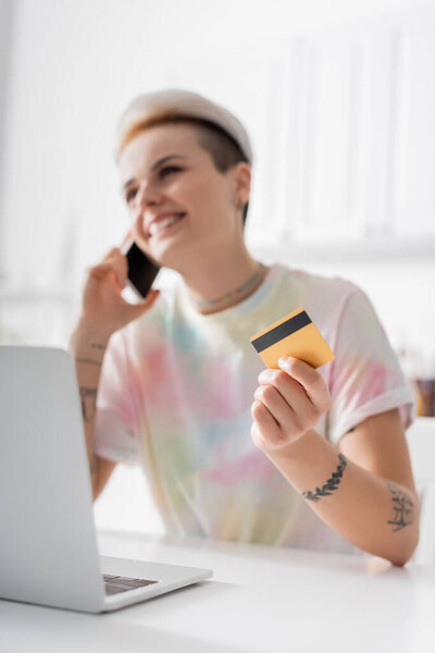 cheerful woman with credit card talking on mobile phone near laptop, blurred background