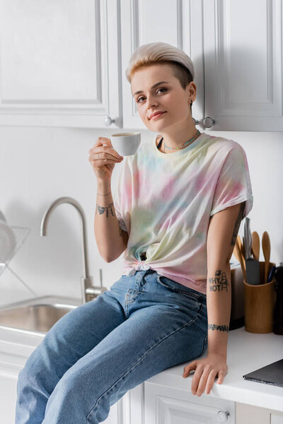 smiling woman sitting on kitchen worktop with coffee cup and looking at camera