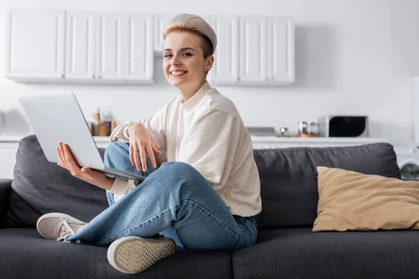 woman with laptop sitting on couch with crossed legs and smiling at camera