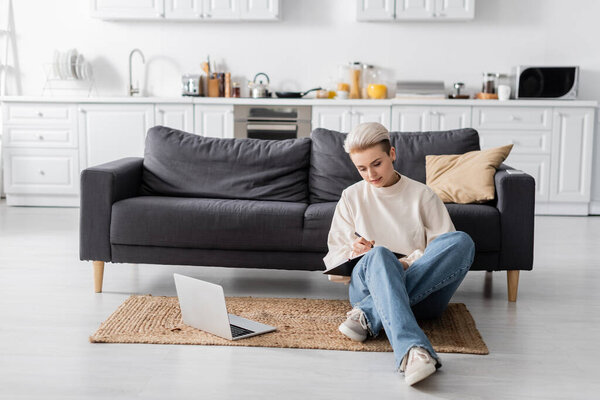 full length of woman in jeans writing in notebook near laptop on floor in spacious open plan kitchen