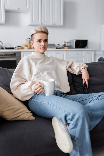 модная женщина в белом свитере держит чашку чая и смотрит кино по телевизору дома