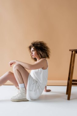 Beyaz elbiseli modaya uygun kıvırcık model bej arka planda sandalyenin yanında oturuyor. 