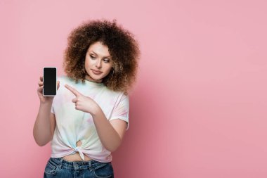 Güzel ve kıvırcık kadın pembe üzerine izole edilmiş cep telefonunu işaret ediyor. 