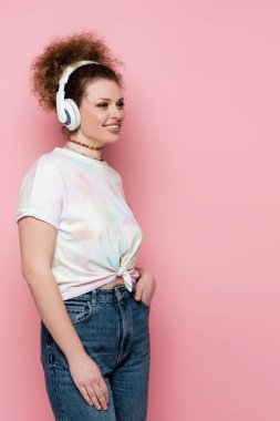 Kot pantolonlu ve tişörtlü mutlu bir kadın pembe renkte izole edilmiş kulaklıkla müzik dinliyor. 
