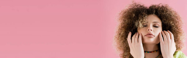 Young curly model with closed eyes touching face isolated on pink, banner